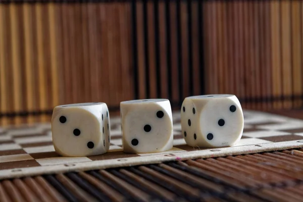 Verem a három fehér műanyag dobókocka barna fa sakktáblát háttér. Kocka hat oldalán fekete pöttyökkel. 3-as szám — Stock Fotó