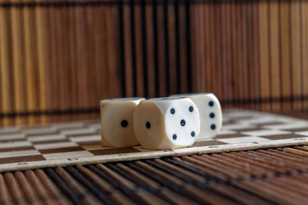 Verem a három fehér műanyag dobókocka barna fa sakktáblát háttér. Kocka hat oldalán fekete pöttyökkel. Rundom száma — Stock Fotó