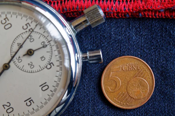 Euro coin with a denomination of 5 euro cents and stopwatch on worn blue jeans with red stripe backdrop - business background