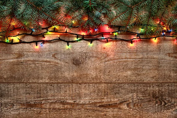 Christmas lights and fir tree branches on wooden background. New Year festive decorations with colorful glowing Christmas lights. Colorful garland on wooden table. Flat lay, view from above — Stock Photo, Image