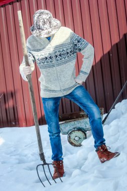 Kazaklı ve kot pantolonlu bir adam bahçede karda duruyor ve elinde bir yaba tutuyor..