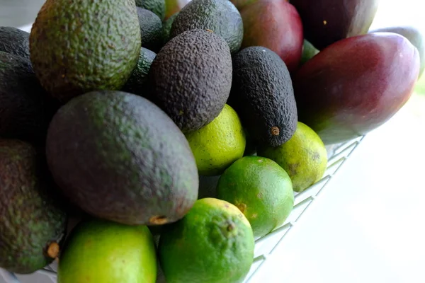 Avokado, mango ve kireç ile beyaz masa üzerinde duran meyve sepeti dolu