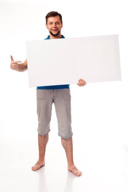 Beyaz bir işareti ile poz sakallı bir adam. Reklam, logo ve diğer yerleştirmek için kullanılabilir. Mavi bir tişört ve gri pantolon giymiş. 