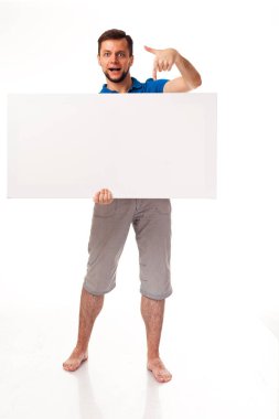 Beyaz bir işareti ile poz sakallı bir adam. Reklam, logo ve diğer yerleştirmek için kullanılabilir. Mavi bir tişört ve gri pantolon giymiş. 