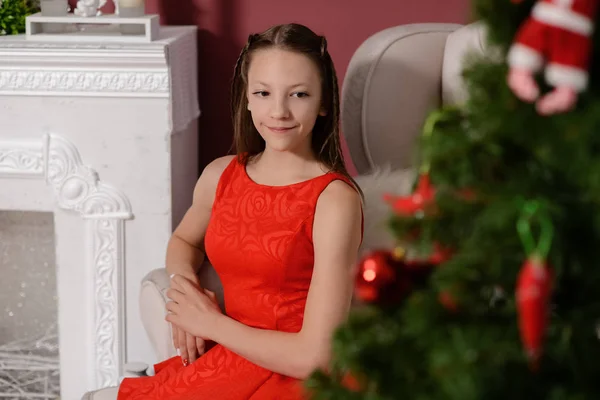La chica en el vestido rojo al lado de la chimenea blanca, decorar un árbol de Navidad con juguetes —  Fotos de Stock