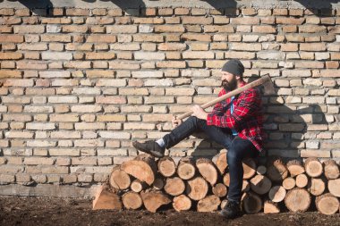 Oduncu ya da oduncu, kırmızı ekose gömlekli, tuğla duvar arkasındaki ağaç gövdelerinde oturuyor..