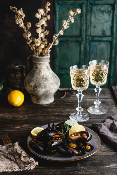 Cooked mussels with shells served on plate with two glasses of white wine on wooden table