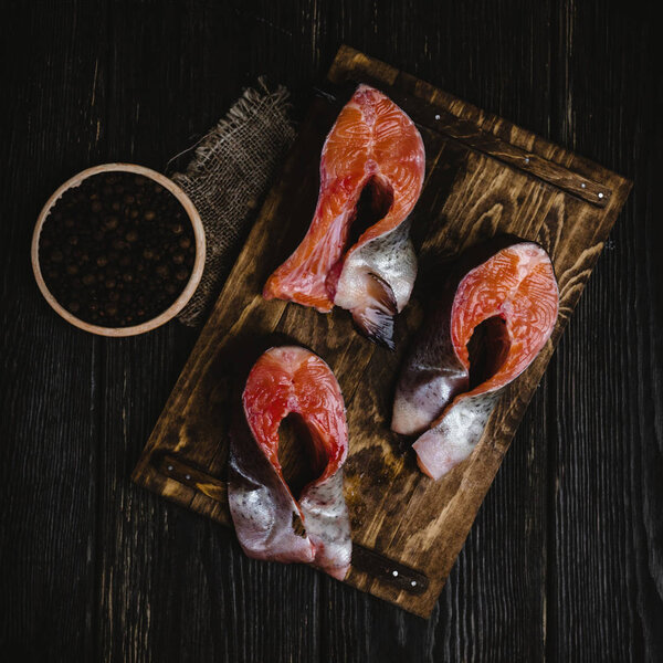 top view of sliced salmon fish on wooden cutting board with sackcloth and peppercorns on rustic table  