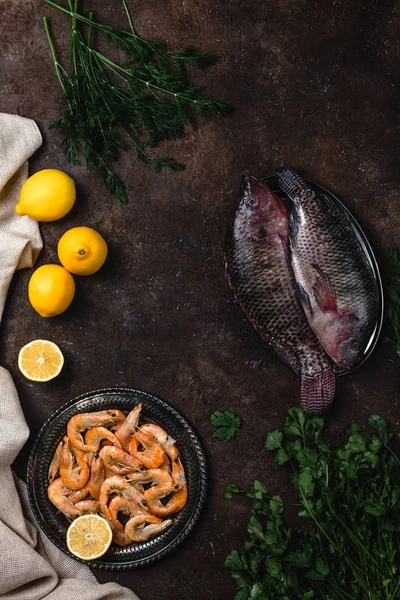 top view of raw fish, shrimp, herbs with lemons and tablecloth on dark table top