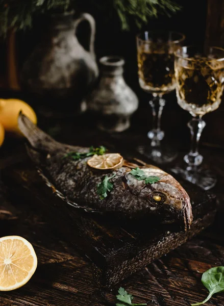 Baked fish with lemon and herbs on wooden board with white wine glasses on dark wooden table — Stock Photo