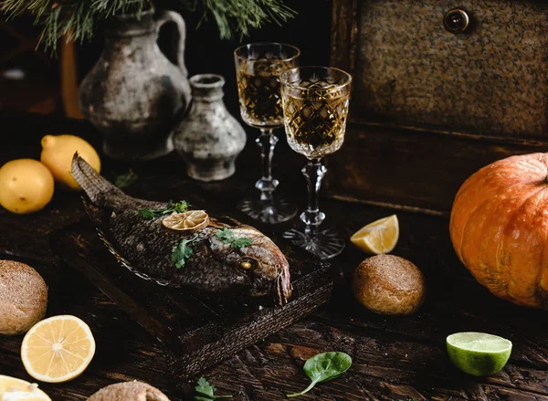 Baked fish with lemon and herbs on wooden board with white wine glasses on dark wooden table — Stock Photo