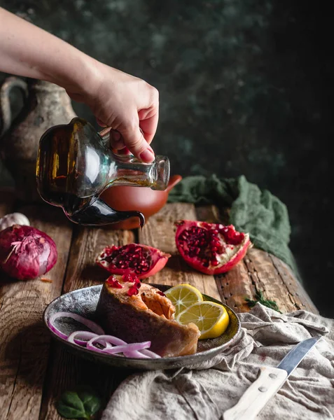 Hembra mano verter aceite sobre filete de salmón al horno con limón y cebolla en el plato con granada en la mesa de madera — Stock Photo