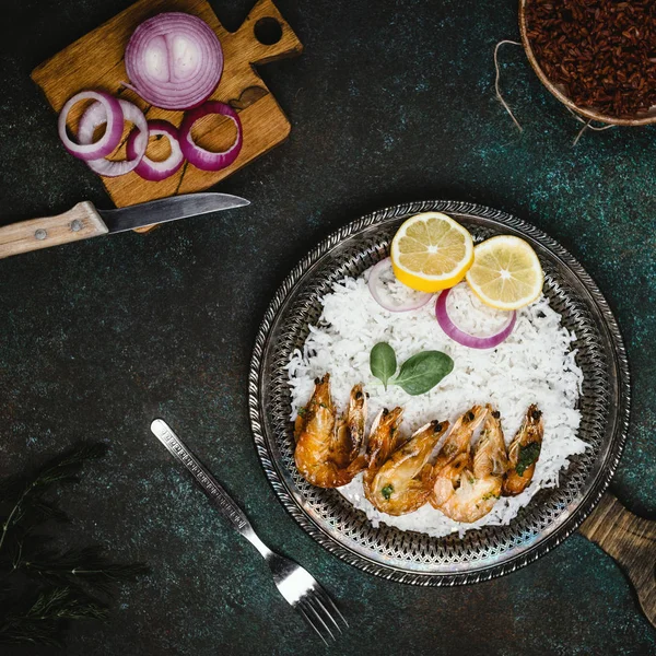 Vista superior de camarones cocidos con limón y cebolla en bandeja de metal rústico con guarnición de arroz en mesa oscura - foto de stock