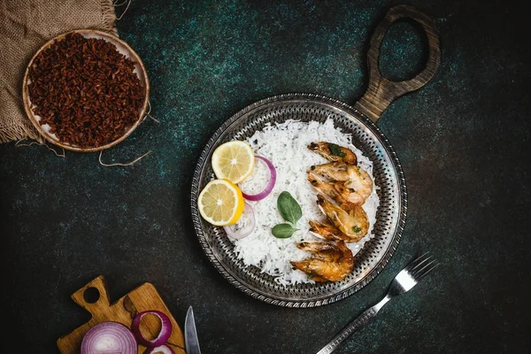 Vista superior de camarões cozidos com limão e cebola na bandeja de metal rústico com prato de arroz na mesa escura — Fotografia de Stock