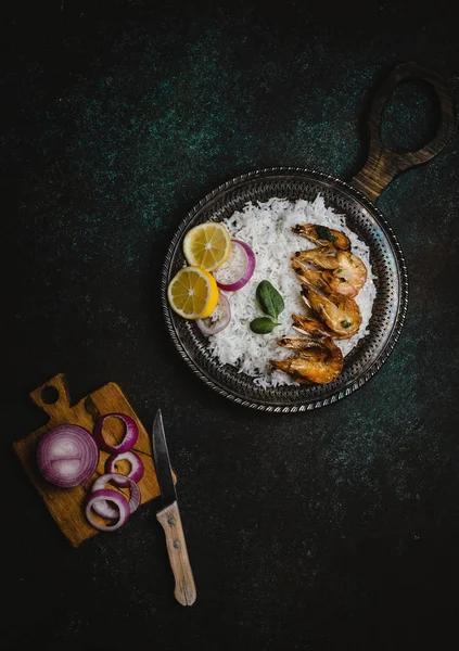 Vista superior de camarões cozidos com limão e cebola na bandeja de metal rústico com prato de arroz na mesa escura — Fotografia de Stock