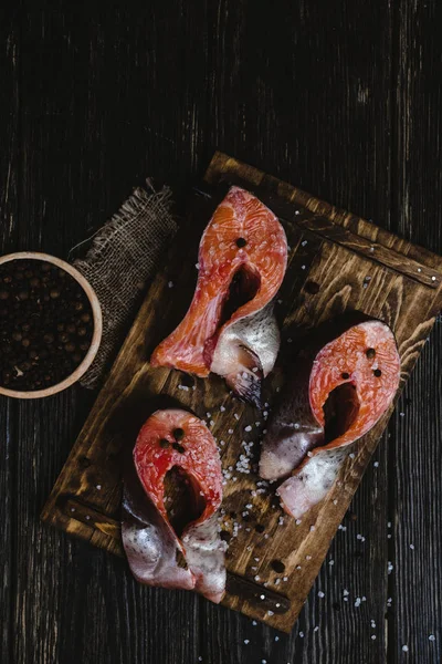 Vue du dessus du saumon frais tranché sur planche à découper en bois avec sac, sel et grains de poivre sur table rustique — Photo de stock