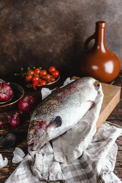 Pescado crudo fresco sobre tabla de cortar de madera y cebollas con ajo y tomates cherry sobre mesa rústica - foto de stock