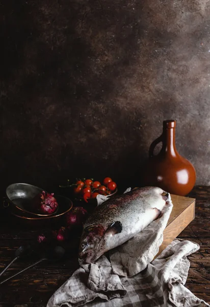 Vista de perto de peixes frescos na tábua de corte de madeira e cebolas com tomates cereja na mesa rústica — Fotografia de Stock