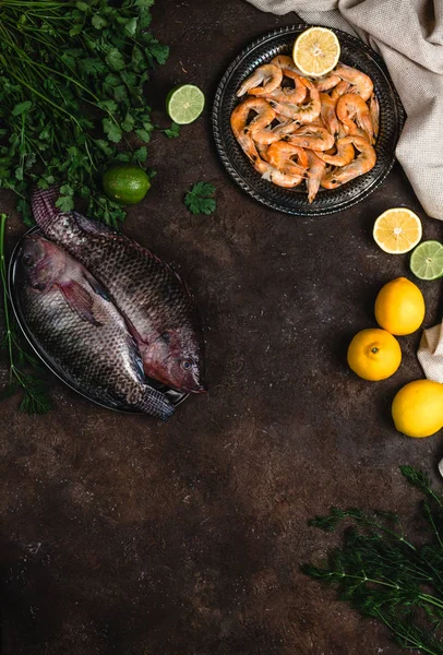 Fresh raw fish, shrimp, herbs with lemons and tablecloth on dark table top — Stock Photo