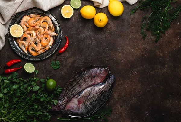 Raw fish, chili peppers, shrimp, herbs with lemons and tablecloth on dark table top — Stock Photo