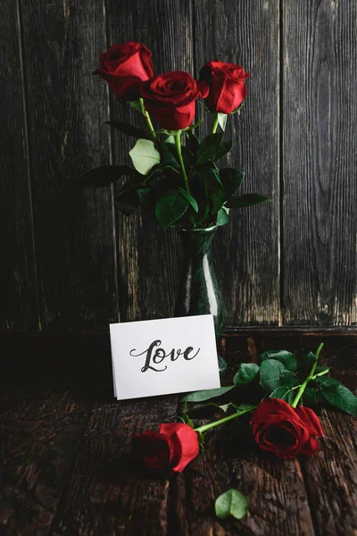 Roses rouges dans un vase et carte de vœux Love sur fond bois minable, concept de Saint Valentin — Photo de stock
