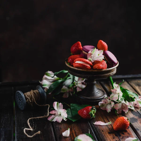Tazón con dulces macarrones y fresas en mesa de madera con flores - foto de stock
