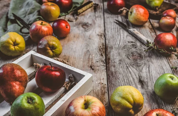 Крупный план яблок, груш, деревянной коробки, ножа, ножниц, чешуек и кухонного полотенца на деревенском столе — стоковое фото