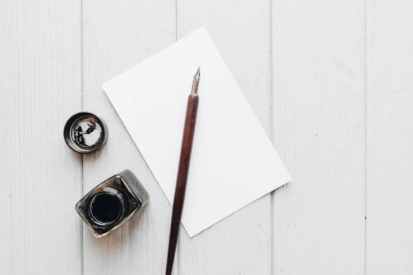 set of vintage dip pen, inkpot and blank paper sheet on white wooden table