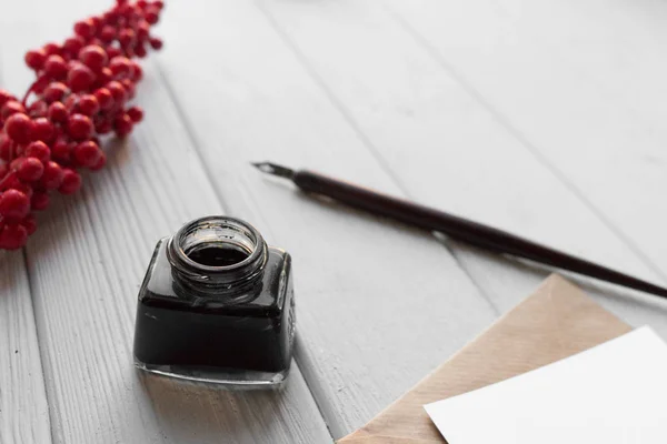 set of vintage dip pen, inkpot, blank paper sheet with envelope and red berries on white wooden table