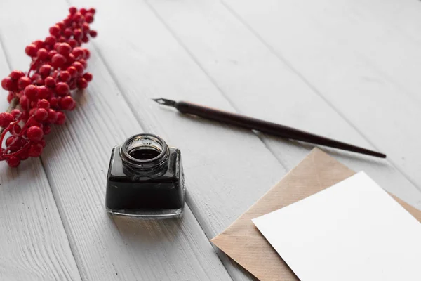 set of vintage dip pen, inkpot, blank paper sheet with envelope and red berries on white wooden table