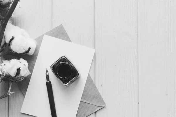 set of vintage dip pen, inkpot, blank paper sheet, envelope and cotton branch on white wooden table, black and white