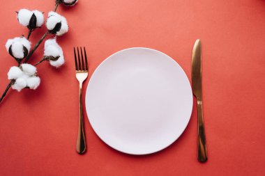 white plate, fork, knife and cotton flowers on red table, elegant tableware 