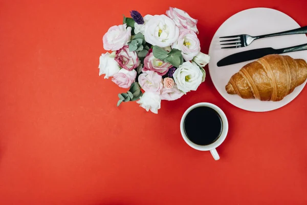 Top view of cup of coffee with flowers and croissant on plate,  red background