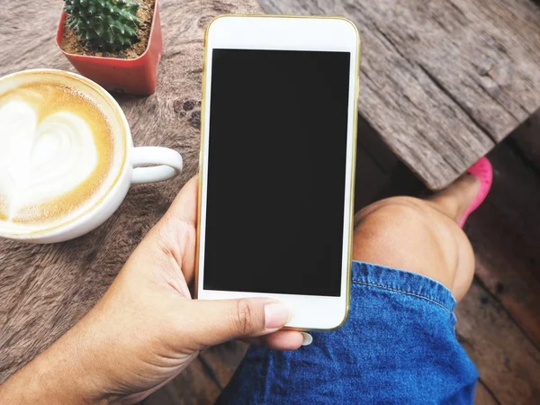 Kahve Kupası ahşap arka plan ile akıllı telefon kullanan kadın