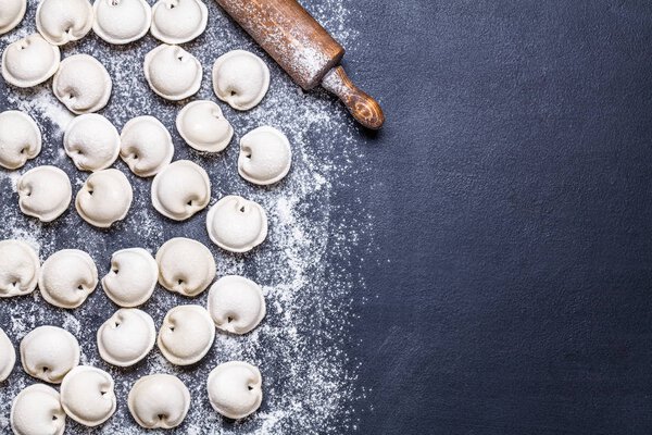 Сырые пельмени на черном фоне, мука, скалка, ложка, горизонтально, плоская лежа
