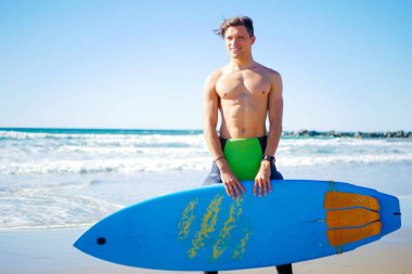 Gömleksiz modeli sörfçü Pasifik okyanus yakın duran mavi sörf tahtası sert top tutan. İyi bir ruh hali, pozitif fotoğraf, yakışıklı supermodel.