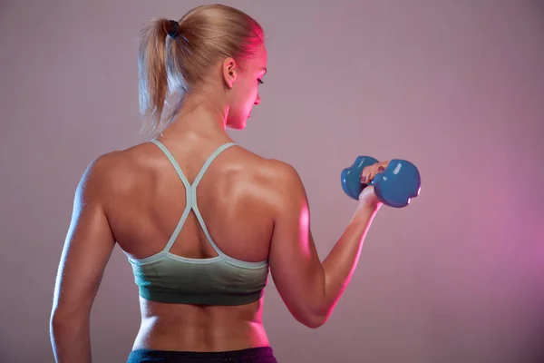 A blond sporty girl holds a dumbbell in her hands, shakes a muscular. Against a dark background — Stock Photo, Image