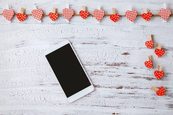 Smartphone ligt op een witte houten tafel met decoratieve rode harten. plek voor tekst. bovenaanzicht — Stockfoto