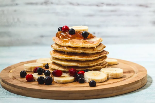 Pfannkuchen mit Honig, Bananen, Marmelade und Beeren auf einem hölzernen Teller auf einer leichten Hintergrundkarte, Restaurantrezeptkonzept. serviert in — Stockfoto