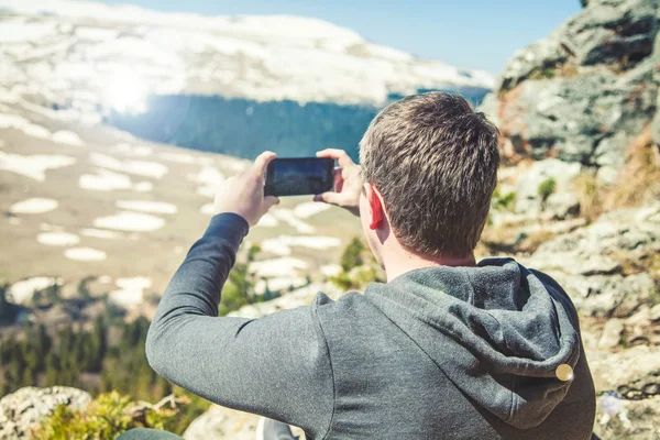 Viagem cara senta-se no topo da montanha e tira fotos do smartphone View from back of the tourist traveler on background mountain — Fotografia de Stock