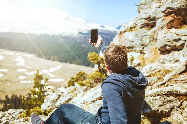 Viagem cara senta-se no topo da montanha e tira fotos do smartphone View from back of the tourist traveler on background mountain — Fotografia de Stock