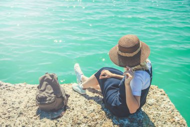 Genç ve güzel kadın turist kız güzel bir mesafe görünüyor güneşli bir deniz kıyısı üzerinde oturur. Gelen turist Gezgin arkasında görüntülemek.