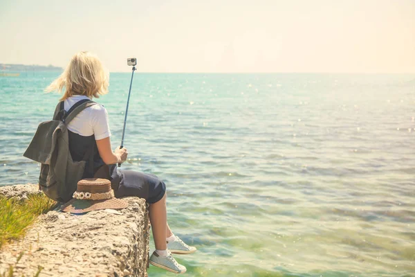 Pretty young woman tourist takes selfie portrait at the seashore on a sunny day Girl takes photo for travel blog. View from back of the tourist traveler