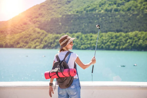 Menina viajante faz selfi com uma câmera de ação em um lago de montanha. Ela faz uma foto para o blog de viagens. Vista de trás do viajante turístico — Fotografia de Stock
