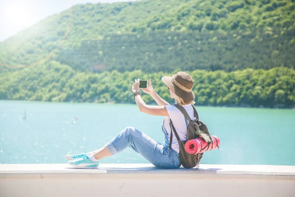 Menina viajante faz selfi com um smartphone em um lago de montanha. Ela faz uma foto para o blog de viagens. Vista de trás do viajante turístico — Fotografia de Stock