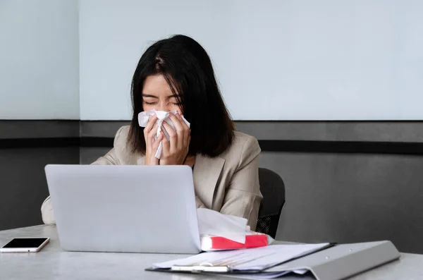 Sick bussinesswoman sitting at office desk with laptop, sneezing and coughing in facial tissue while working in meeting room. Spreading and risk of coronavirus covid-19 in workplace