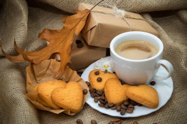 bir fincan sütlü kahve, bir kalp, kaba kumaş arka plan üzerinde bir hediye şeklinde kurabiye ile natürmort