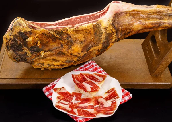 Closeup of serrano ham leg with plate and tablecloth over black — Stock Photo, Image