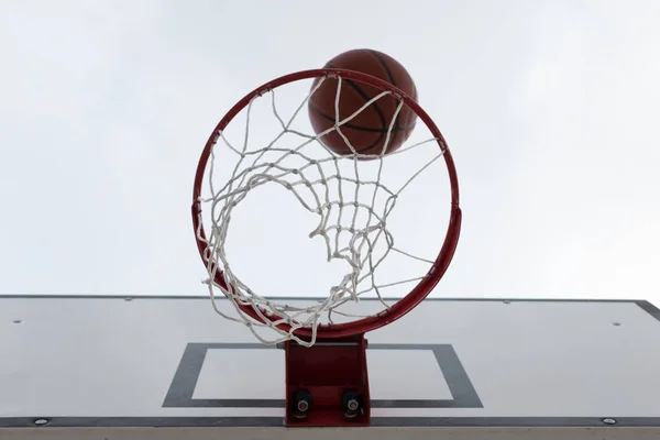 Ball inside red basketball hoop, basket against white sky. Outdoor basketball court. White board.