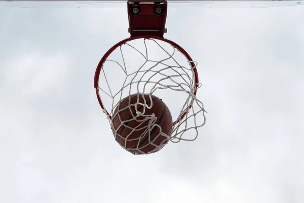 Orange ball inside red basketball hoop, basket against white sky. Outdoor basketball court.